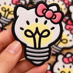 May include: An embroidered patch featuring a white cat character head shaped like a lightbulb. The design includes a bright yellow glow, a pink bow on the right ear, and a silver-grey threaded base, all outlined with a thick black border.