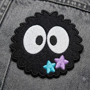 May include: A circular embroidered patch featuring a fuzzy black creature with two large white oval eyes and black pupils. The design includes a small light blue star and a small light purple star positioned near the bottom right edge of the patch.