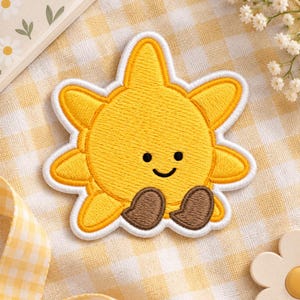 May include: A yellow embroidered sun patch with a white border, featuring a simple black stitched face with two small eyes and a curved mouth, and two brown stitched feet positioned at the bottom of the circular sun shape.