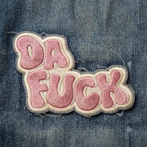 May include: An embroidered patch featuring the text DA FUCK in bubble-style pink lettering with a cream-colored fill and a grey border. The patch is stitched onto a blue denim fabric background with visible frayed threads along the edges.