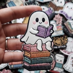 May include: An embroidered patch featuring a white ghost with pink cheeks sitting on a stack of four colorful books while reading a purple book. The ghost has a simple black-lined face and is holding the open purple book with both hands.