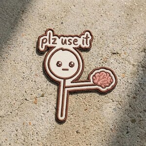 May include: An embroidered patch featuring a white magnifying glass with a brown border and a cartoon face. The text "plz use it" is embroidered in brown above the magnifying glass, and a pink brain is attached to the handle by a thin line.