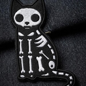 May include: An embroidered patch featuring a black cat silhouette with a white stitched skeleton design. The cat has a skull-shaped head with large eye sockets, a small nose, and a visible rib cage, spine, and tail bone structure in white thread.