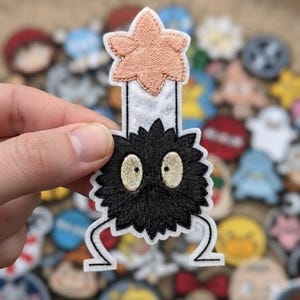 May include: An embroidered patch featuring a black, spiky, circular creature with two large, pale yellow eyes, holding a white candle topped with a light peach-colored flame. The patch has a white border and a white backing material.