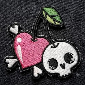 May include: An embroidered patch featuring a pink heart and a white skull connected by a green leaf stem. The heart is accented with white highlights and crossed white bones behind it, all outlined in black thread on a dark fabric background.
