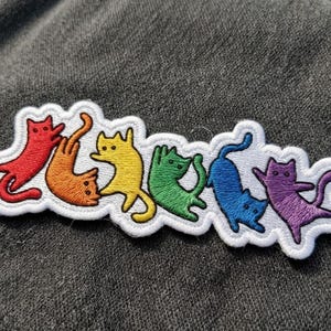 May include: An embroidered patch featuring a row of six cats in a rainbow color sequence of red, orange, yellow, green, blue, and purple. Each cat is depicted in a playful, stretched-out pose with a white stitched border outlining the entire shape.