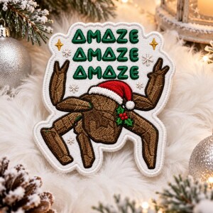 May include: An embroidered patch featuring a brown, multi-legged creature wearing a red and white Santa hat with a holly sprig. The text AMAZE is stitched in green three times above the creature, accented by small gold stars and white snowflakes.