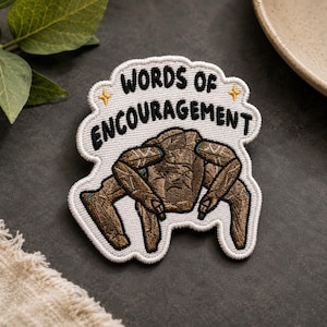 May include: An embroidered patch with a white background and a scalloped edge. It features the black text "WORDS OF ENCOURAGEMENT" above a brown, textured, four-legged creature illustration with two small yellow star icons on either side.