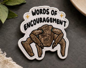 Può includere: Una toppa ricamata con sfondo bianco e bordo smerlato. Presenta il testo nero "WORDS OF ENCOURAGEMENT" sopra l'illustrazione di una creatura marrone a quattro zampe con texture e due piccole stelle gialle su entrambi i lati.