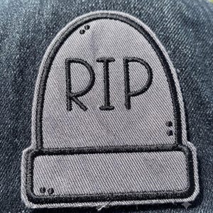 May include: A gray tombstone-shaped embroidered patch featuring the black stitched text RIP. The patch has a thick black border and is attached to a dark blue denim fabric background. The design includes small black stitched dots as accents.