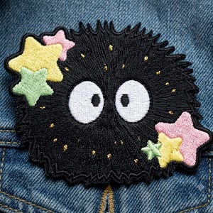 May include: An embroidered patch featuring a round, fuzzy black creature with two large white oval eyes. The creature is accented with small gold metallic threads and surrounded by several pastel-colored stars in shades of yellow, pink, and light green.