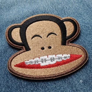 May include: An embroidered patch featuring a tan monkey face with black outlines, large ears, and a wide red mouth showing white teeth with silver metal braces. The design is stitched with a textured thread finish on a blue denim fabric background.