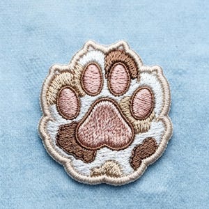 May include: An embroidered patch shaped like a cat paw print. The design features a large central pad and four smaller toe pads in shades of dusty pink, surrounded by white, tan, and dark brown fur-like stitching with a beige border.