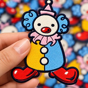 May include: An embroidered clown patch featuring a cream-colored face, a red nose, and blue curly hair. The clown wears a pink ruffled collar, a yellow and blue body with two white buttons, and large red shoes, all outlined in dark navy blue thread.