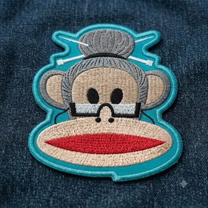 May include: An embroidered patch featuring a sock monkey with gray fur, a tan face, and a bright red mouth. The monkey wears black-rimmed glasses and has a gray bun hairstyle accented with two white knitting needles, all set against a teal border.