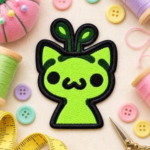 May include: An embroidered patch featuring a bright lime green cat-like creature with two antenna-like sprouts on its head, black circular eyes, and a black mouth, outlined in thick black thread against a neutral fabric background.