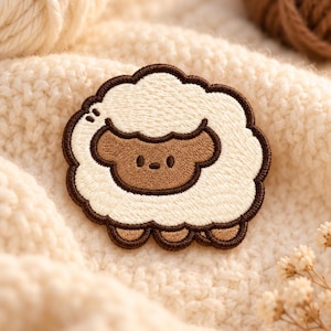 May include: An embroidered patch featuring a stylized sheep with a brown face and cream-colored wool. The design is outlined in dark brown thread, showcasing a simple facial expression with two small eyes and a horizontal line for a mouth.