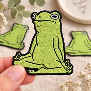 May include: An embroidered patch featuring a light green frog sitting in a meditative lotus position with black outlines. The frog has large white eyes with black pupils and is making a mudra hand gesture. The patch has a thick black border.