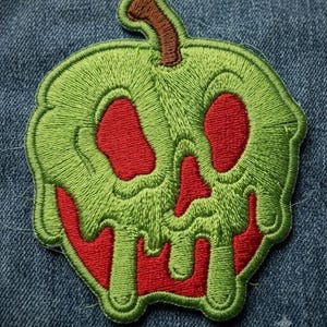 May include: An embroidered patch shaped like a red apple covered in dripping green slime, featuring a brown stem at the top. The design includes two hollow eye sockets and a nose opening, revealing the red base color beneath the green embroidery.
