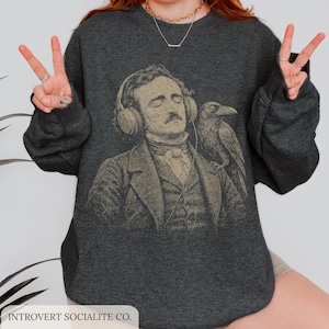 Può includere: Una felpa girocollo grigio antracite scuro con una stampa grafica beige di Edgar Allan Poe che indossa cuffie con un corvo appollaiato sulla spalla. Il design include un cavo che si estende dalle cuffie. Il testo INTROVERT SOCIALITE CO. è stampato nell'angolo in basso a sinistra.