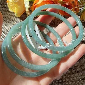 May include: Three smooth, circular jade bangles in a pale green hue. Each bangle features a polished, translucent surface with subtle natural variations in colour. A small white paper tag with the text ITEM: is attached to one of the bracelets.