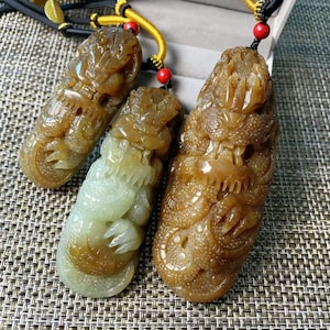 May include: Three carved stone pendants featuring intricate dragon motifs. Each pendant is suspended from a black cord with a yellow accent thread and a small red bead. The stones exhibit varying shades of amber, brown, and pale green, with detailed textures depicting the dragon's scales and features.