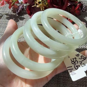 Op de afbeelding: Een stapel van vier gladde, bleek doorschijnende groene jade armbanden met een afgerond profiel. Aan de armbanden is een klein wit rechthoekig label bevestigd met de handgeschreven tekst #59, 58.5 en 11.4 in zwarte inkt.
