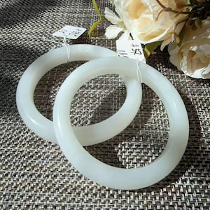 May include: Two smooth, opaque white circular bangles with a polished finish. Each bangle features a small white paper tag attached with a thin white string. The tags contain handwritten black text. The bangles are positioned near cream flowers.