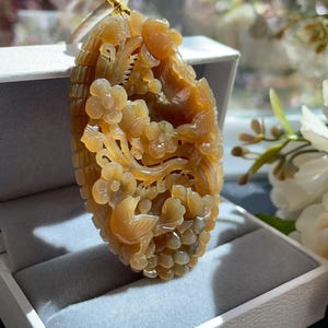 Könnte beinhalten: Ein ovaler, bernsteinfarbener Anhänger mit detaillierten Reliefschnitzereien von blühenden Blumen und einem kleinen Vogel. Der durchscheinende Stein zeigt einen warmen, goldbraunen Farbton mit feinen Texturen in den Blumen- und Vogelmotiven.