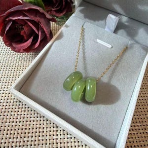 May include: A gold-toned chain necklace featuring three smooth, translucent light green circular jade-like beads strung together. The necklace is presented inside a light grey velvet-lined jewellery box, showcasing a polished and minimalist design.