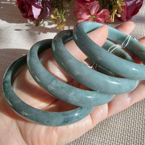 May include: A set of four smooth, rounded jade bangles in a muted sage green color. Each bangle features a polished, glossy finish with subtle natural variations in the stone's texture and color, showcasing a solid, circular, and seamless design.