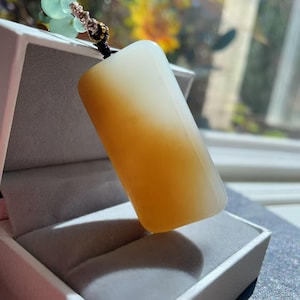 May include: A rectangular pendant featuring a smooth, polished stone with a gradient transition from a warm, translucent amber-yellow at the base to a pale, milky white at the top. The pendant is suspended by a dark cord with small green beads.