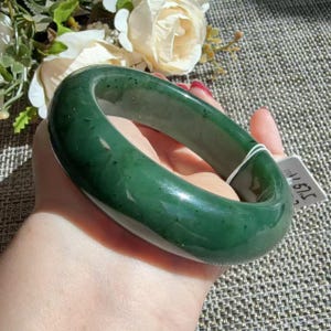 Può includere: Un bracciale rigido in giada verde scuro, liscio e levigato, con profilo arrotondato e sottili inclusioni naturali. Un piccolo cartellino rettangolare bianco è attaccato al lato del bracciale, con il testo stampato 57.5 in inchiostro nero.