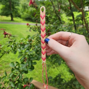 May include: A woven friendship bracelet featuring a chevron pattern in shades of red, orange, and cream. The bracelet has a loop at the top and two braided ties at the bottom, showcasing a detailed knotted design with a consistent color gradient.