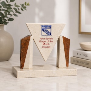 May include: A triangular stone award trophy featuring the New York Rangers logo and the text John Spears Player of the Month January. The piece is constructed from cream-colored stone with dark brown wood accents on the sides and a rectangular base.