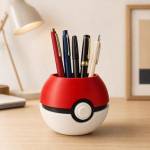 Peut inclure: Un organisateur de bureau sphérique conçu comme une balle rouge et blanche avec une bande centrale noire et un bouton blanc. Cinq stylos de différentes couleurs, dont le rouge, le bleu, le noir et le crème, sont placés verticalement à l'intérieur.