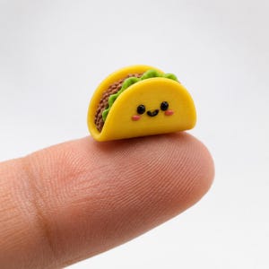 Könnte beinhalten: Eine Miniatur-Taco-Figur in Gelb mit einem lächelnden Gesicht, bestehend aus schwarzen Augen, einem kleinen geschwungenen Mund und rosa Wangen. Die Taco-Schale ist leuchtend gelb und enthält eine braune, texturierte Füllung sowie grünen Salat.