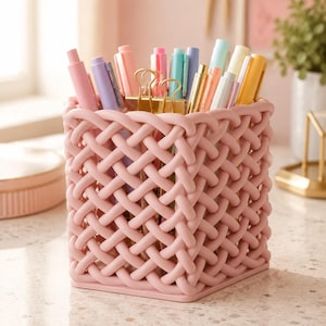 Peut inclure: Un porte-stylo carré rose clair avec un design extérieur tressé en forme de treillis. Le récipient contient divers stylos dans des tons de rose, violet, bleu et jaune, ainsi que deux pinces à dessin en métal doré insérées à l'intérieur.