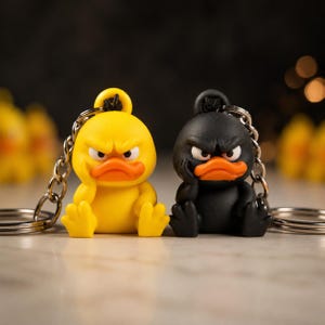 Peut inclure: Deux porte-clés représentant des figurines de canard de dessin animé, l'un jaune vif et l'autre noir mat. Les deux canards ont un bec orange, les sourcils froncés et une main posée sur la joue, attachés à un anneau en métal argenté.
