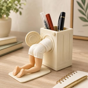 May include: A cream-colored desk organizer shaped like a washing machine with a figure in white shorts kneeling and reaching into the open door. The top of the unit serves as a container for three pens, including one clear, one red, and one black pen.