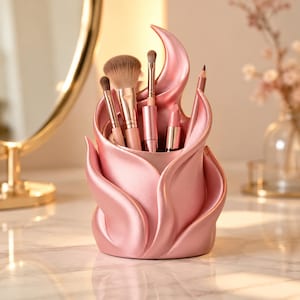 Op de afbeelding: Een metallic roze make-up kwastenhouder in de vorm van een gestileerde bloem met gebogen, overlappende bloemblaadjes. De houder bevat diverse make-up kwasten, een lippenstift en een potlood voor een georganiseerde opslag van cosmetica.