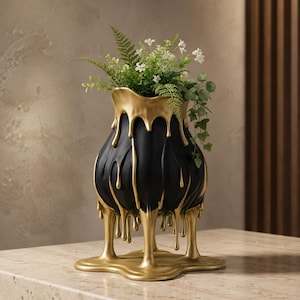 May include: A matte black vase with a rounded body and a wide opening, featuring a decorative gold-colored drip effect that cascades from the rim down the sides and pools at the base. The vase contains a cluster of green ferns and white flowers.