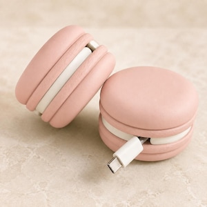 Puede incluir: Dos organizadores de cables redondos de color rosa claro con forma de macarrón y una banda central blanca. Uno está colocado en posición vertical, mientras que el otro está plano con un cable de carga USB-C blanco que sale por el lateral.