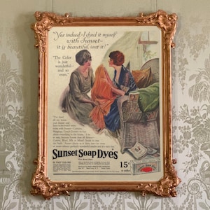Könnte beinhalten: Eine Vintage-Werbung für Sunset Soap Dyes in einem verzierten kupferfarbenen Rahmen. Der Druck zeigt zwei Personen, die orangefarbenen Stoff halten, mit dem Text: Yes indeed-I dyed it myself with Sunset-it is beautiful, isnt it! und 15 cents a cake.