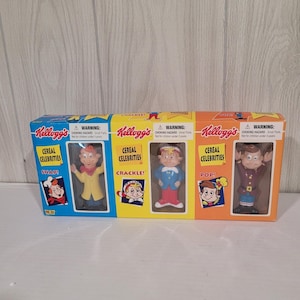 Może przedstawiać: Zestaw trzech figurek Kellogg's Cereal Celebrities przedstawiający Snap, Crackle i Pop w prostokątnym pudełku. Opakowanie jest podzielone na niebieskie, żółte i pomarańczowe sekcje, z których każda zawiera nazwę postaci oraz etykietę ostrzegawczą.