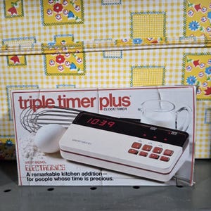 May include: A West Bend Triple Timer Plus clock and timer in a white and black rectangular housing with red digital display and red buttons. The packaging features text including triple timer plus, West Bend, and electronics on a white background.