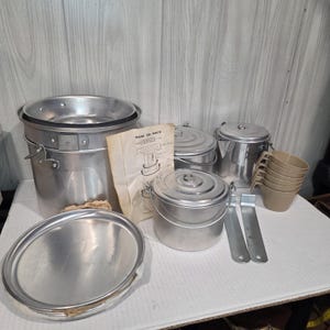 以下が含まれることがあります： 銀色のアルミニウム製キャンプ用調理器具セット。蓋付きの大きな鍋、蓋付きの小さな鍋、背の高い容器、5つのベージュのプラスチック製カップ、2つの金属製ハンドルツール、および「How to Pack」と書かれた説明書が含まれています。
