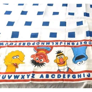 May include: A white fabric sheet featuring a repeating pattern of blue squares. A horizontal red stripe contains the alphabet in blue letters. Below this, illustrations of Big Bird, Elmo, Bert, and Cookie Monster appear above a second red stripe.