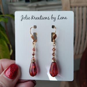 May include: A pair of dangle earrings with a teardrop-shaped red and orange glass bead and gold accents. The earrings are on a white card with the text "Jolie Kreations by Lena".