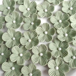 May include: A collection of sage green ceramic flower embellishments with white pearl centers. The flowers have detailed petal textures and are arranged closely together, creating a decorative floral pattern. These are ideal for craft projects.